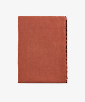 A folded rust linen Nico Tablecloth with a thin decorative stitch along one edge, set against a plain white background. This hand-finished detail enhances the tablecloth’s elegant simplicity.