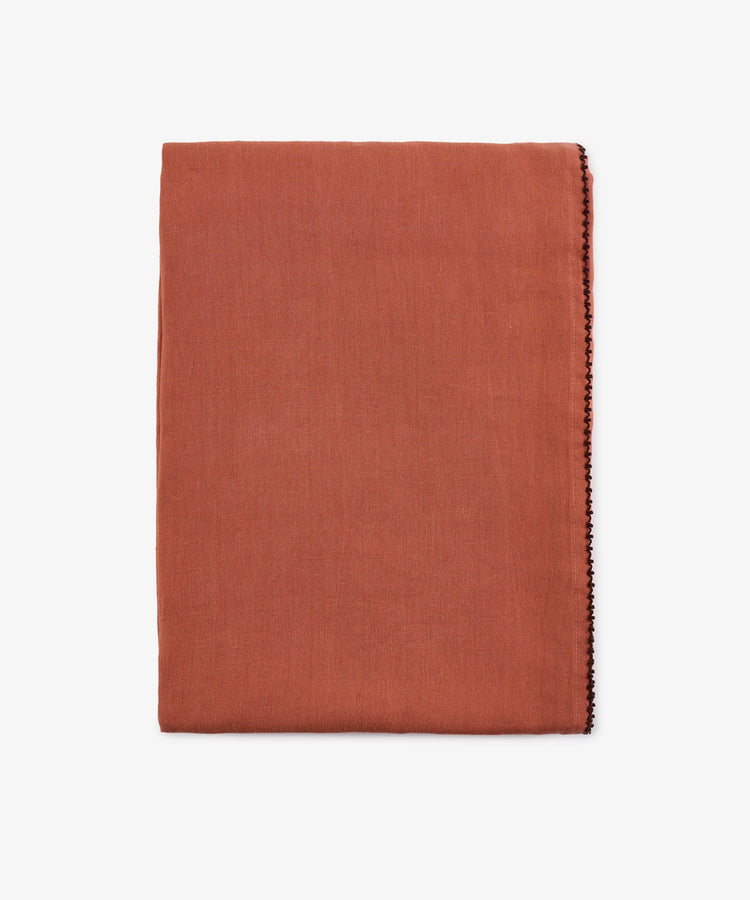 A folded rust linen Nico Tablecloth with a thin decorative stitch along one edge, set against a plain white background. This hand-finished detail enhances the tablecloth’s elegant simplicity.