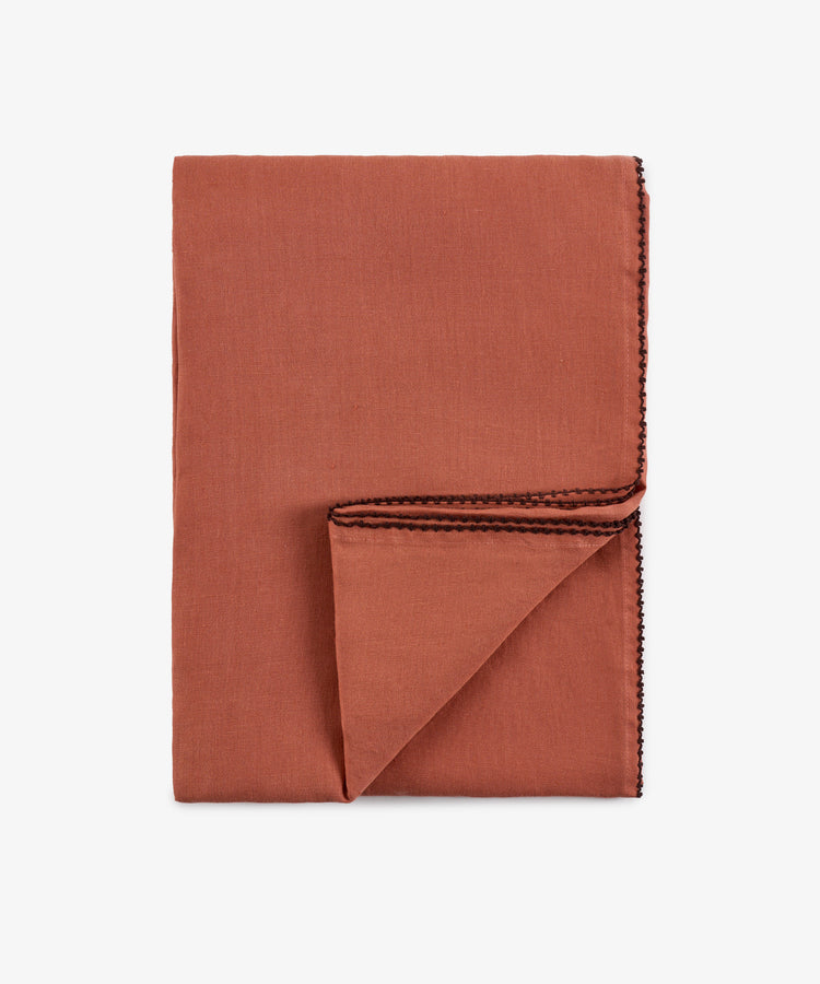 A folded rust-colored Nico Tablecloth with black stitched trim, hand-finished for superior quality, displayed on a white background.