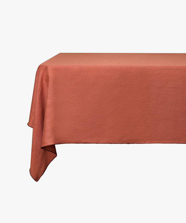 A rectangular table covered with the Nico Tablecloth in plain rust linen, set against a white background.