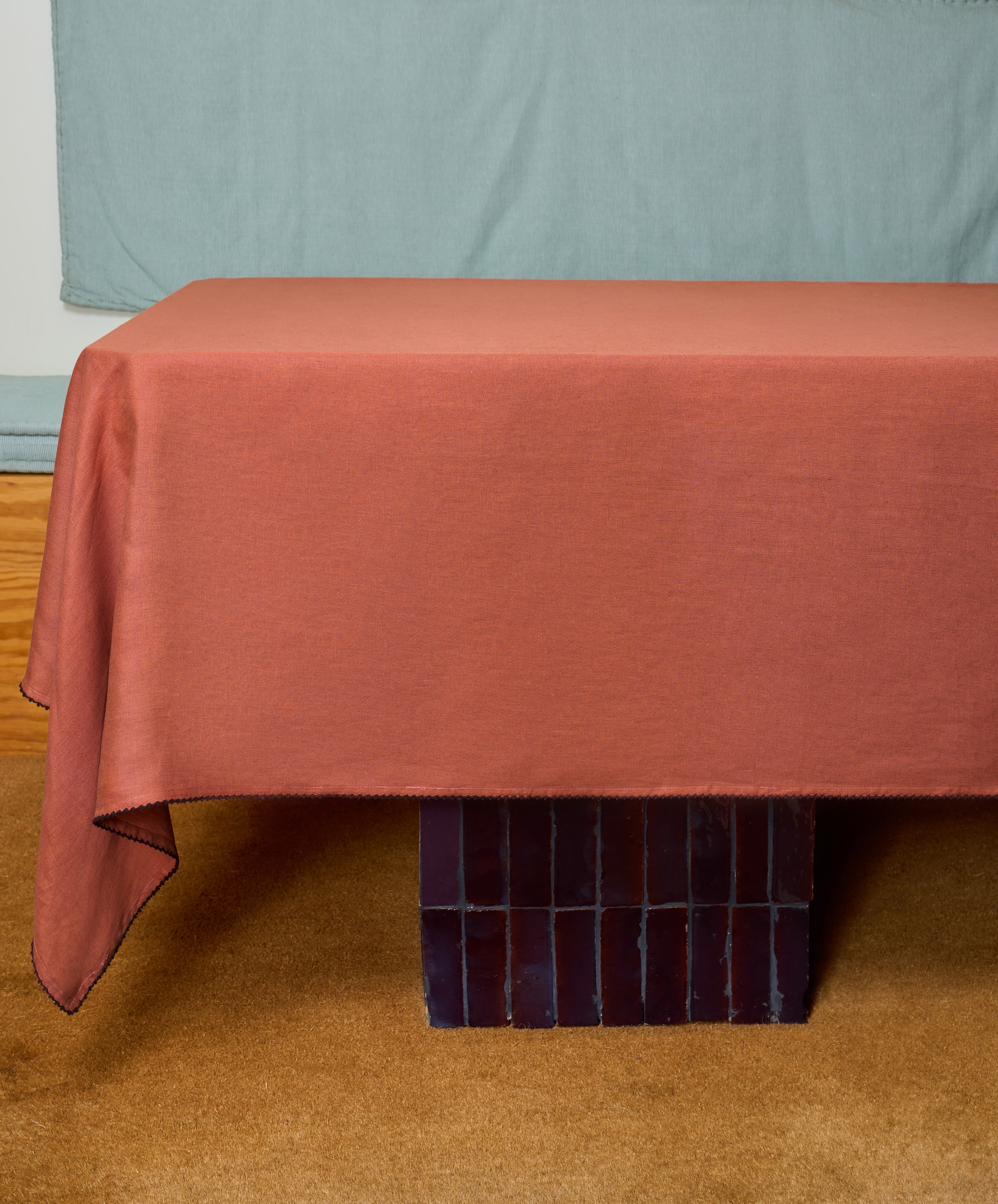 A rectangular table draped with the Nico Tablecloth in hand-finished rust linen stands on a dark block base, set against a light blue background and tan carpet.