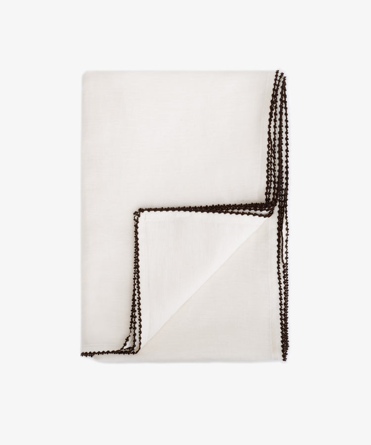 The Nico Tablecloth is a white linen tablecloth with dark contrast stitching along the edges, shown against a plain white backgroundâ€”an elegant choice for table decor.