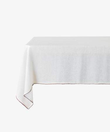 The Nico Tablecloth, made from white linen with subtle red contrast stitching, is elegantly draped over a table and set against a plain white background.