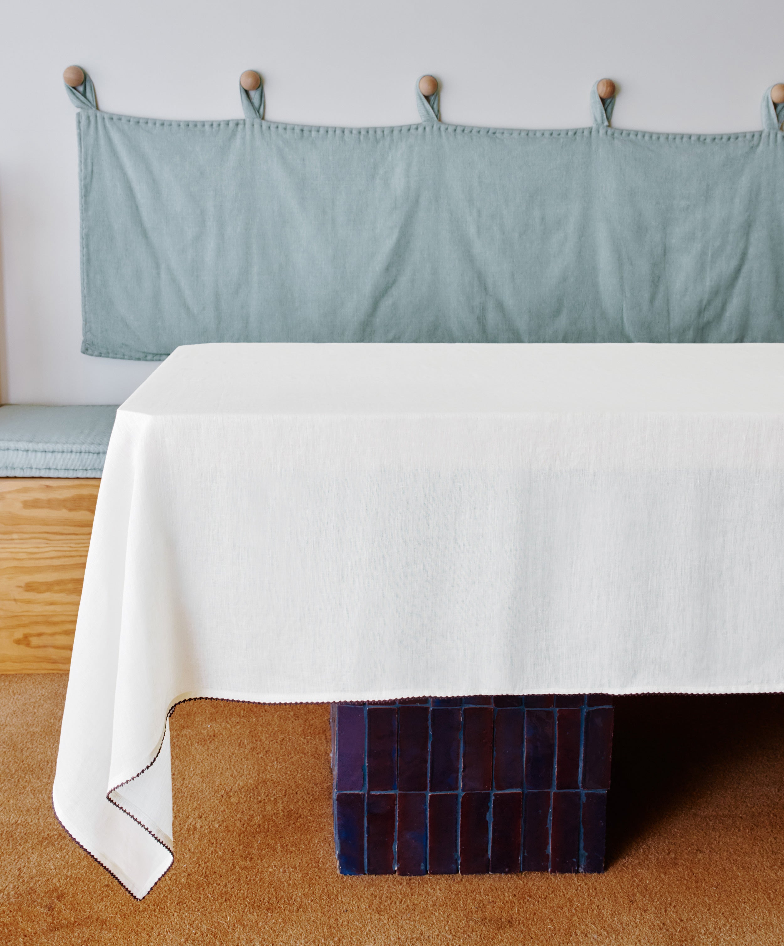 The Nico Tablecloth, made of linen, covers a rectangular table with a blue tiled base; behind it sits a wooden bench with a light blue padded backrest and contrast stitching against a white wall.