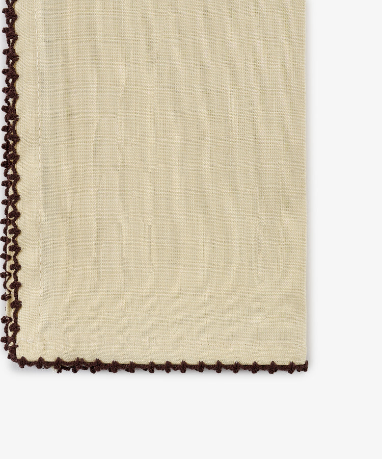 A close-up of the Nico Napkin, Set of 6, in a beige linen fabric with dark brown crochet trim along the left and bottom edges, featuring antique-inspired espresso contrast stitch details.