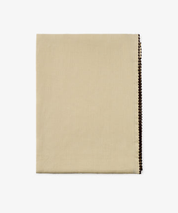 The Nico Tablecloth, a folded beige fabric with a decorative dark edge, is displayed on a white background.