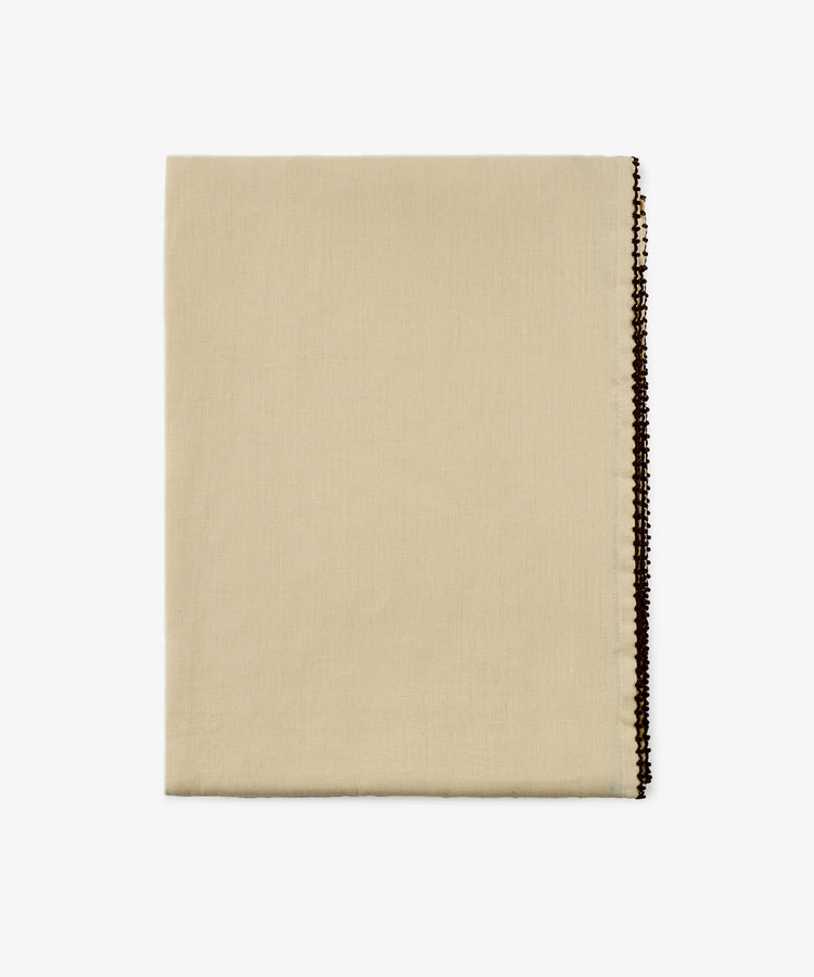 The Nico Tablecloth, a folded beige fabric with a decorative dark edge, is displayed on a white background.