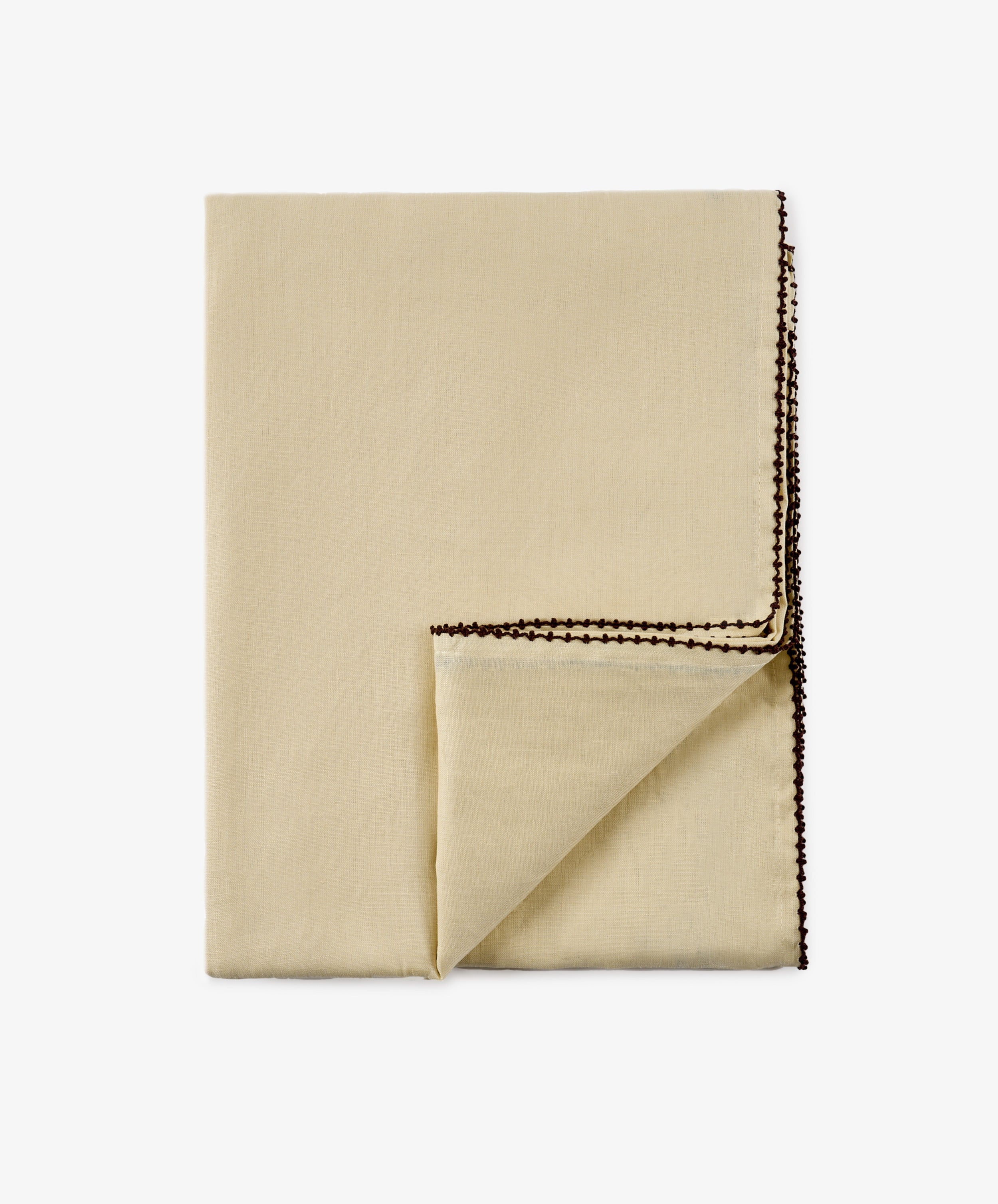 The Nico Tablecloth, crafted from folded beige linen with a hand-finished touch, features a decorative brown stitched edge and is shown with one corner turned over against a plain white background.