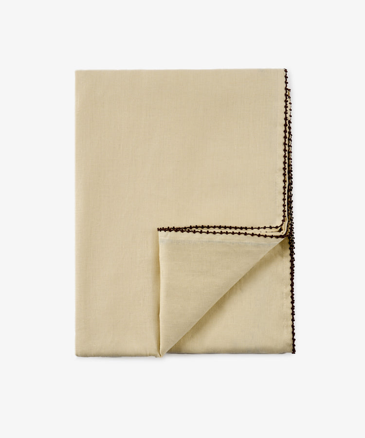 The Nico Tablecloth, crafted from folded beige linen with a hand-finished touch, features a decorative brown stitched edge and is shown with one corner turned over against a plain white background.