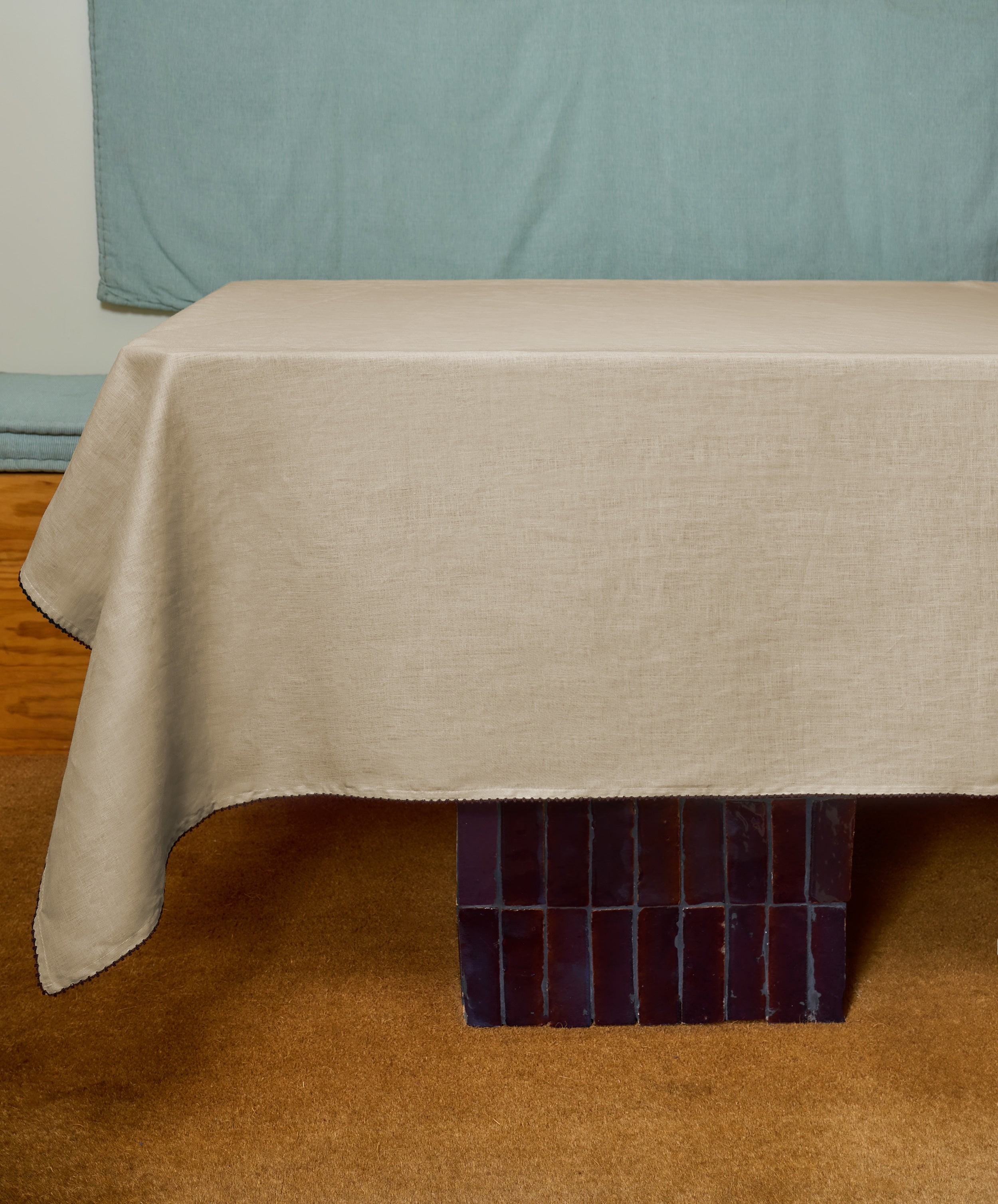 A hand-finished linen tablecloth in antique yellow covers a rectangular table with dark tiles, set on a brown carpet with a light blue backdrop.