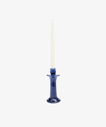 A tall, white taper candle stands in the elegant, glossy Angelita Candleholder, a handcrafted dark blue ceramic piece, against a plain white background.