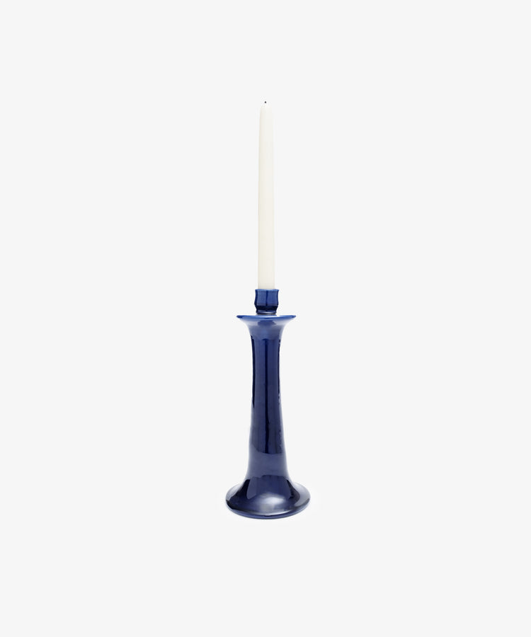 A white taper candle stands upright in the Maria Candleholder, a glossy, dark blue handcrafted ceramic piece, set against a plain white background.