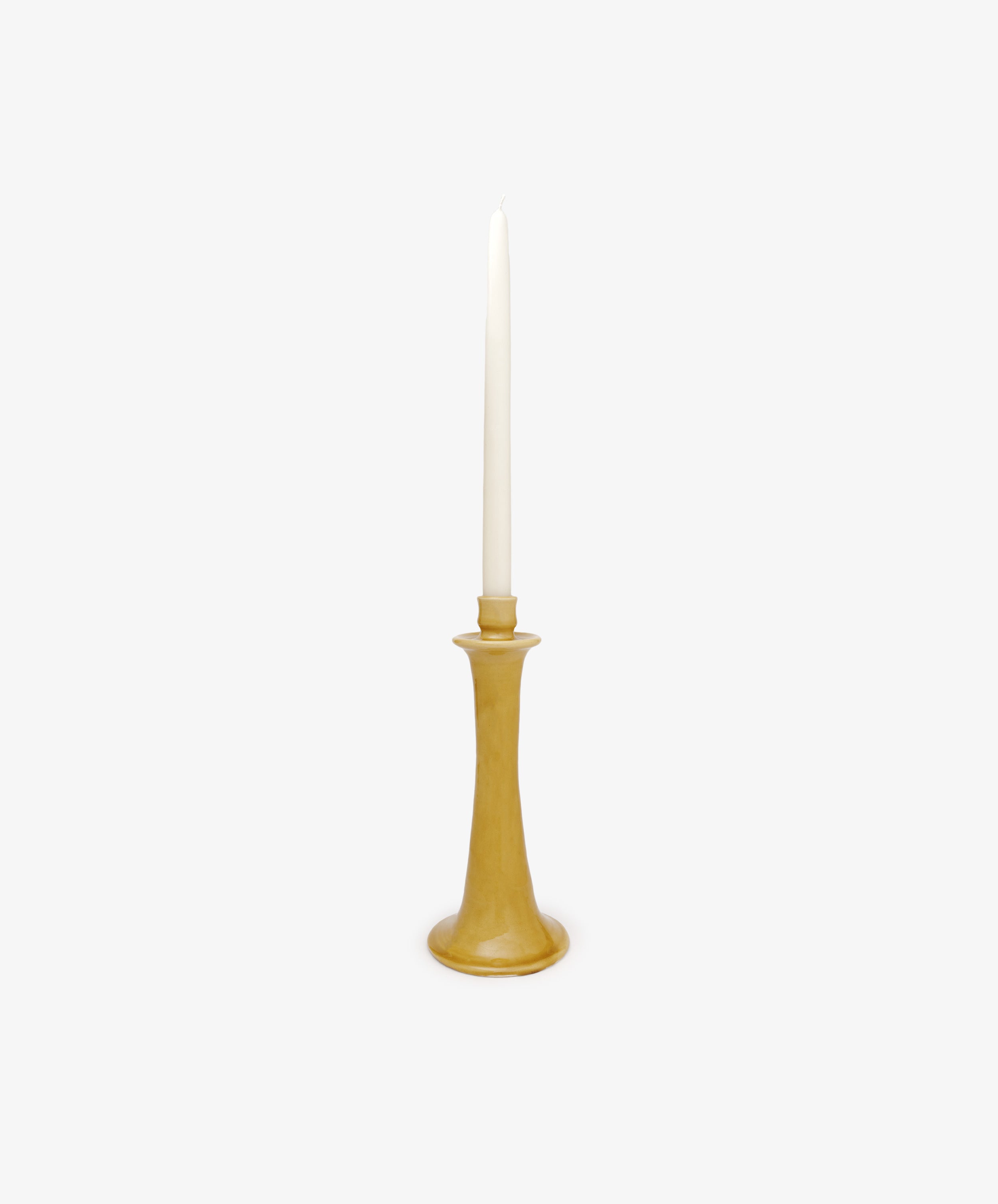 A tall, unlit white taper candle sits in the Maria Candleholder, a simple, handcrafted yellow ceramic holder from Spain, shown against a plain white background.