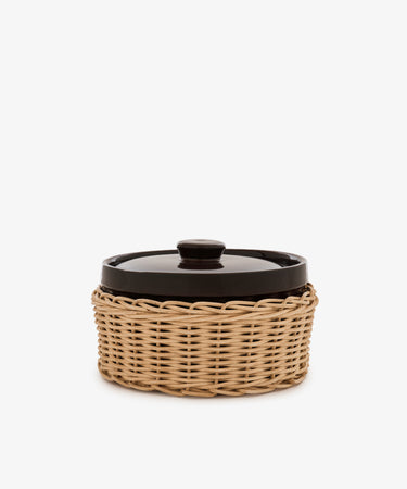 Amany Casserole, a black-lidded ceramic baking dish nestled in a round woven rattan basket, photographed against a plain white background.
