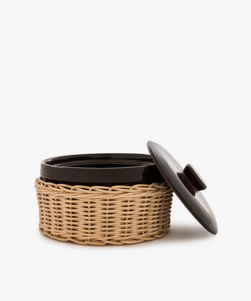 The Amany Casserole, a round ceramic baking dish with a dark lid partially open, sits inside a light-colored woven basket holder against a white background.