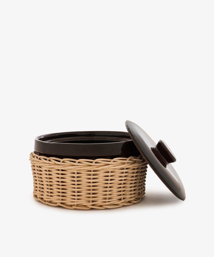 The Amany Casserole, a round ceramic baking dish with a dark lid partially open, sits inside a light-colored woven basket holder against a white background.