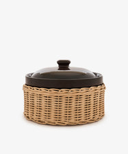 The Amany Casserole, a dark stoneware dish with a lid, is presented inside a light-colored woven wicker basket against a plain white background.