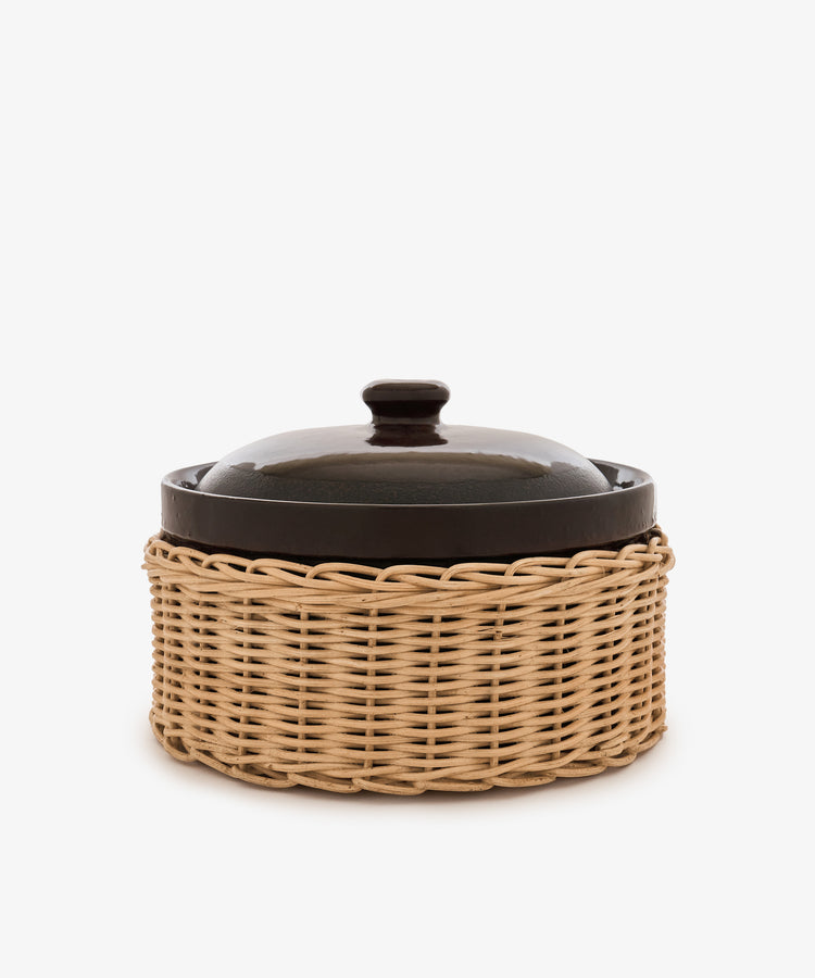 The Amany Casserole, a dark stoneware dish with a lid, is presented inside a light-colored woven wicker basket against a plain white background.