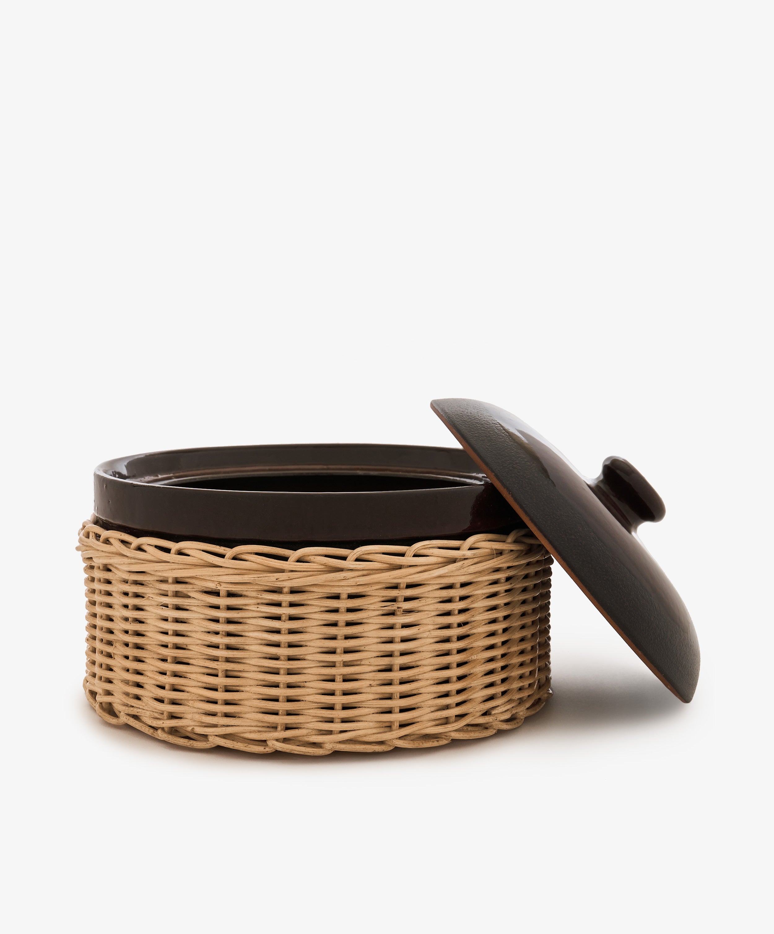 The Amany Casserole, a dark ceramic baking dish with a matching lid partially open, sits in a woven wicker holder against a white background.