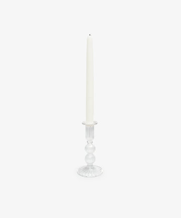 A white taper candle stands upright in the Elsa Candleholder, its clear glass adding an elegant touch to home decor against a plain white background.