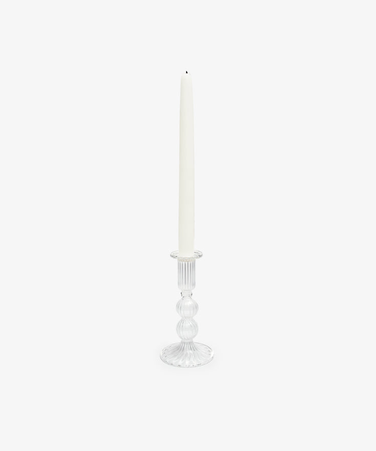 A white taper candle stands upright in the Elsa Candleholder, its clear glass adding an elegant touch to home decor against a plain white background.