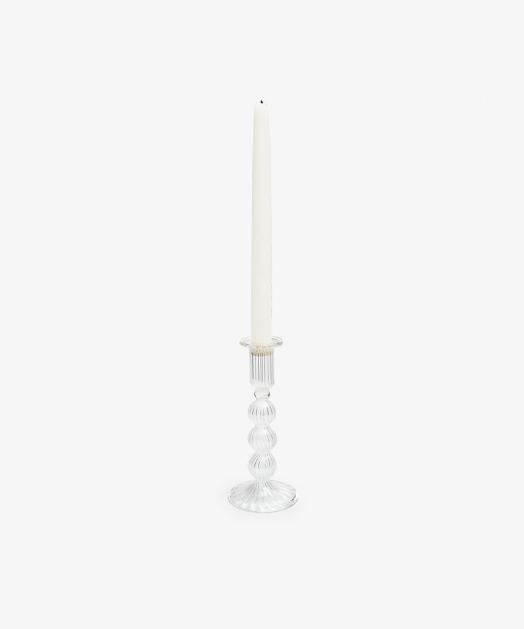 A tall white taper candle sits in the Elsa Candleholder, a clear hand-blown glass piece featuring a ribbed, rounded design, against a plain white background.
