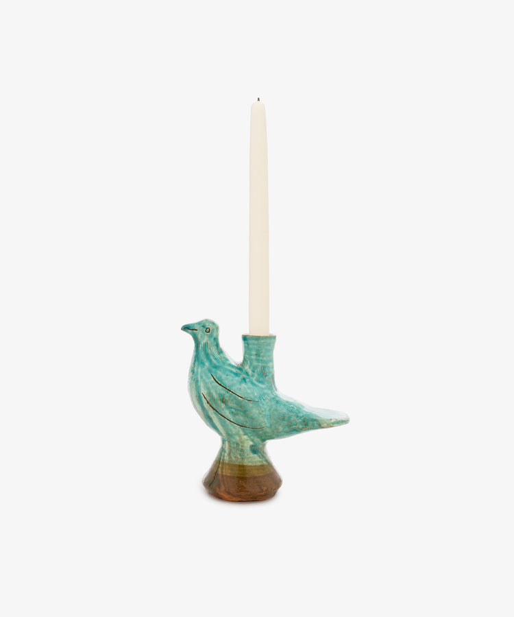 The Peace Dove Candle Holder showcases a ceramic bird gracefully poised atop a decorative pedestal, finished in a tranquil blend of turquoise, sage green, and deep brown glaze. This elegant piece holds a tall white taper candle and stands out beautifully against a plain white background.
