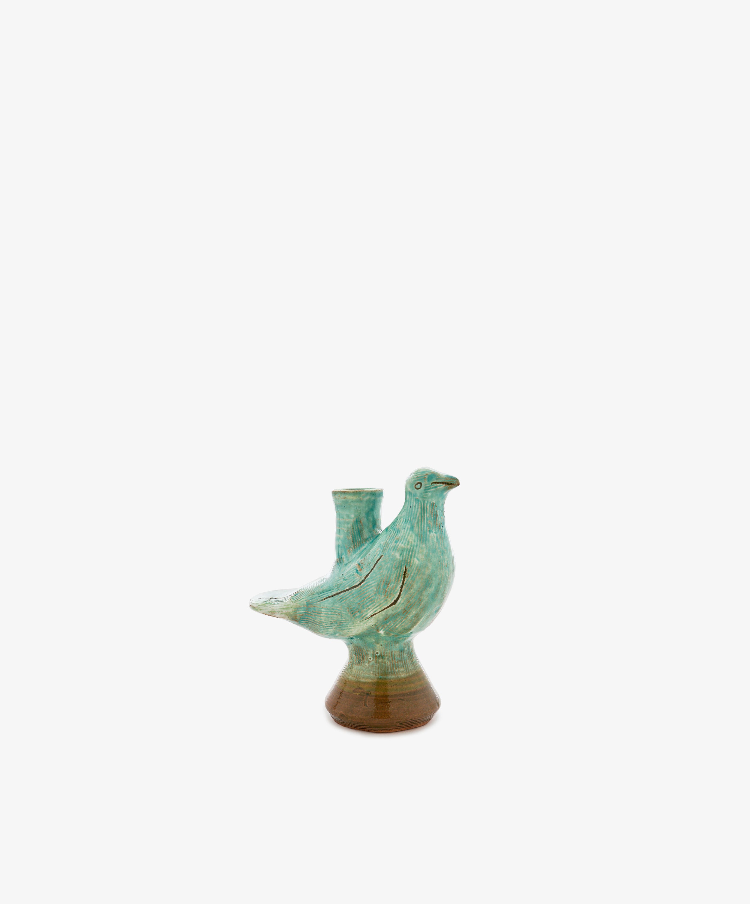 The Peace Dove Candle Holder features a ceramic vessel shaped like a bird, finished in a sage green glaze that highlights its elegant form. A small cylindrical opening on its back makes it perfect for holding a candle, creating an ideal accent piece for a decorative pedestal or shelf. Set against a plain white background, this unique holder is both functional and stylish.