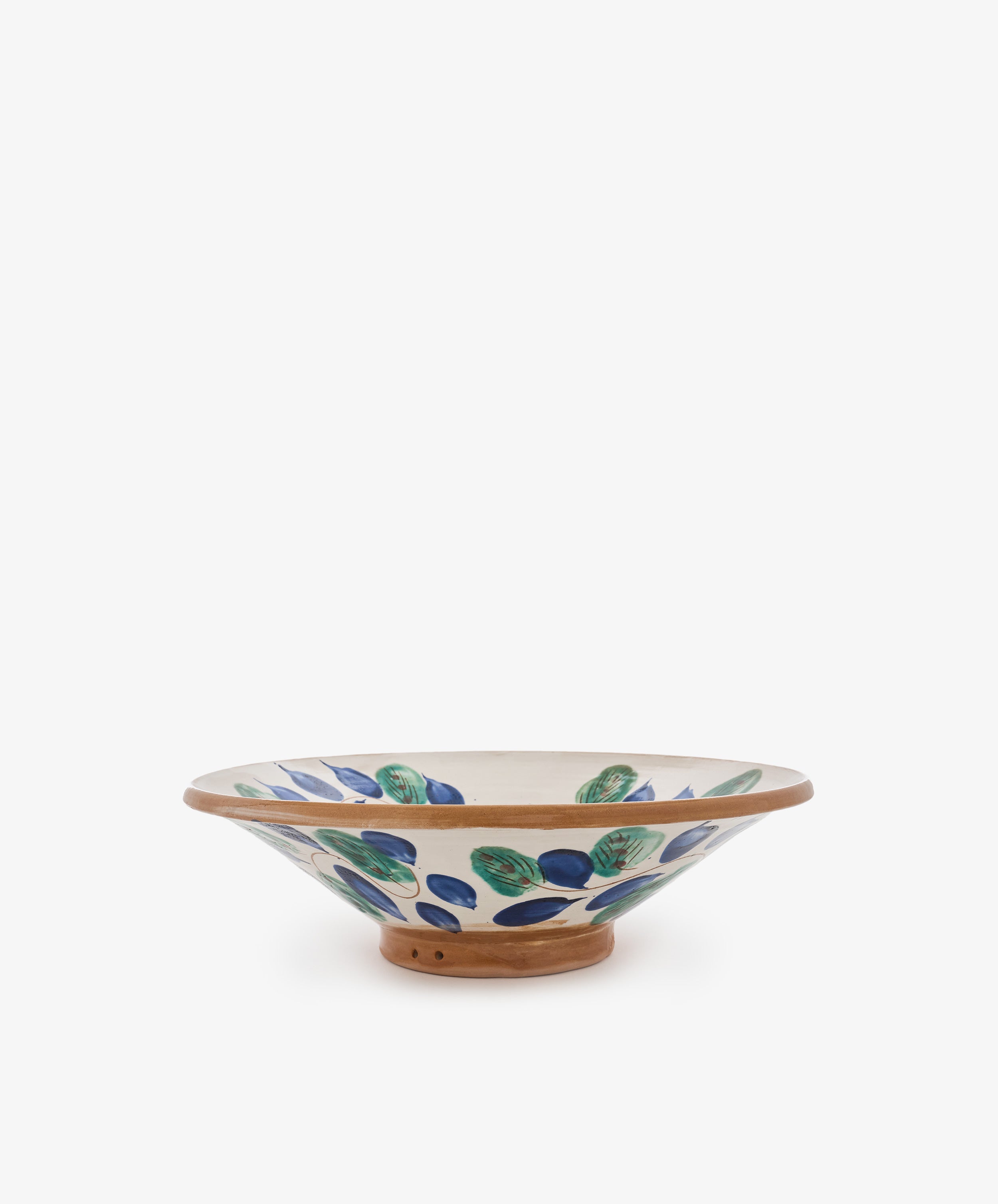 The Midnight Garden Serving Bowl is a shallow, hand-thrown ceramic bowl with a wide rim, inspired by the tradition of Fayoum pottery. It features green and blue leaf patterns on a white background and is finished with a natural brown base.