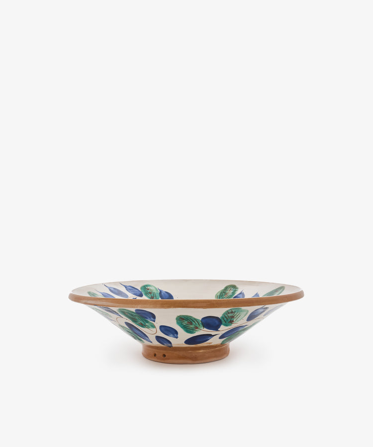 The Midnight Garden Serving Bowl is a shallow, hand-thrown ceramic bowl with a wide rim, inspired by the tradition of Fayoum pottery. It features green and blue leaf patterns on a white background and is finished with a natural brown base.