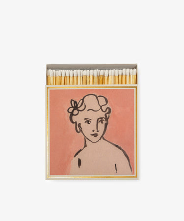Luxury Matches come in an open matchbox with a pink cover, featuring a letterpress-printed line drawing of a person’s bust—curly hair, flowers, and a neutral expression—making them a unique keepsake gift.