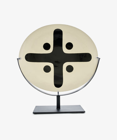 Motif 5, Oval is a round, cream-colored glazed earthenware piece featuring a bold black cross and four dots, displayed upright on a rectangular-based metal stand.