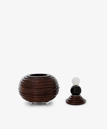 The Curiosity Box, a round, ridged African wood container with three metal feet, is displayed beside its matching lid featuring a black and white spherical handle.