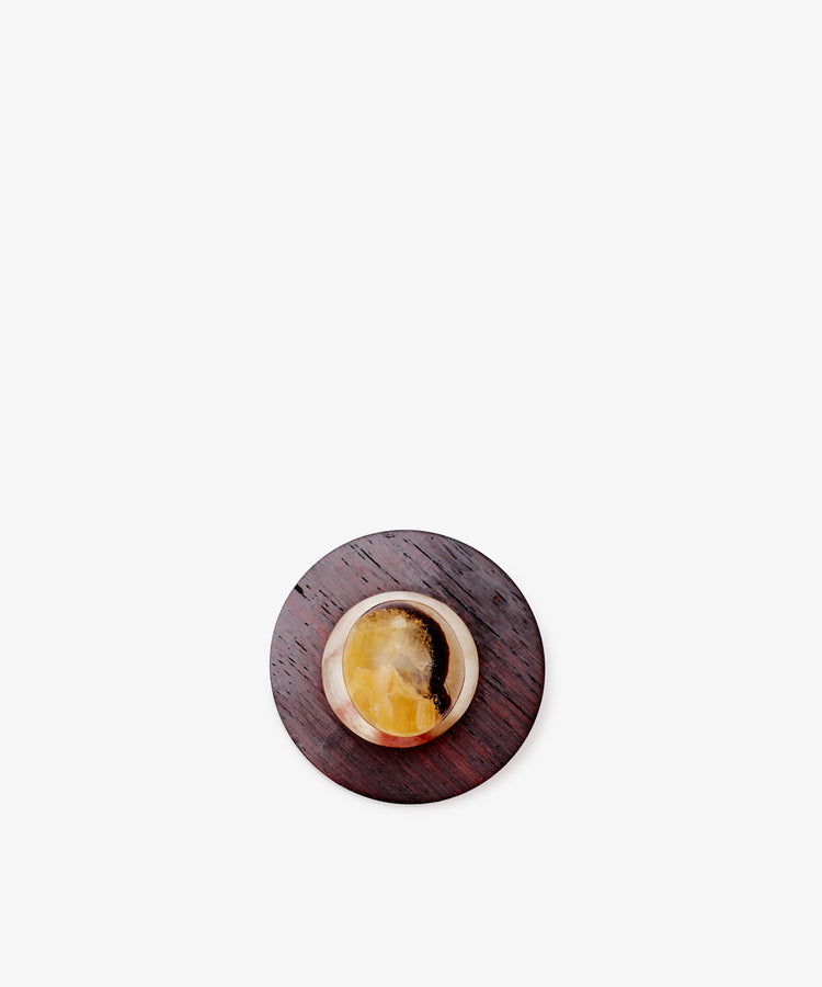 The Curiosity Box—a small, round stone in brown and yellow tones on a dark wooden base—evokes an African wood box, displayed against a plain white background.