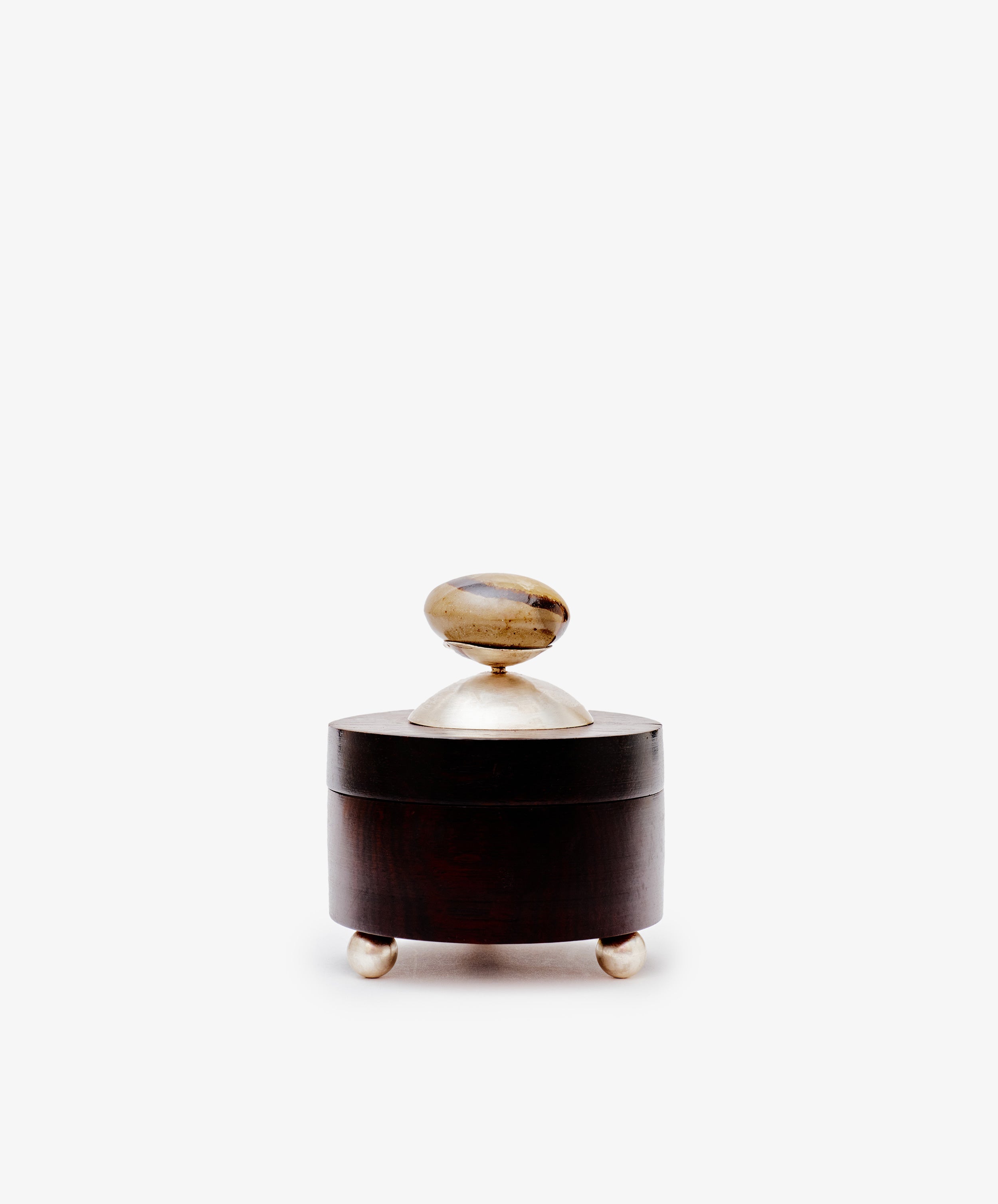 The Curiosity Box is a small, round wooden artisan box by Antje Weber. It includes a lid with a metallic knob, stands on three metallic feet, and is displayed against a plain white background.