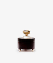 The Curiosity Box is a small, round wooden artisan box by Antje Weber. It includes a lid with a metallic knob, stands on three metallic feet, and is displayed against a plain white background.
