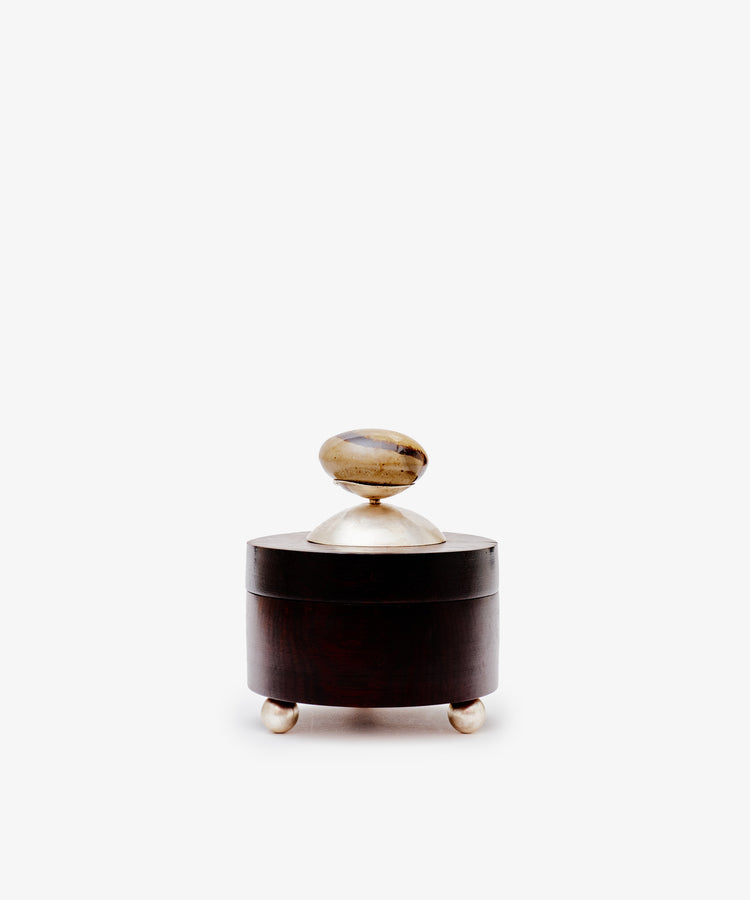The Curiosity Box is a small, round wooden artisan box by Antje Weber. It includes a lid with a metallic knob, stands on three metallic feet, and is displayed against a plain white background.