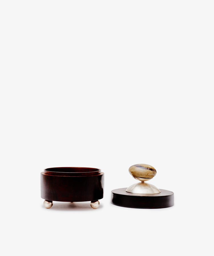 The Curiosity Box is a round African wood box with three small metal feet and a matching lid topped with a stone handle, shown against a white background.