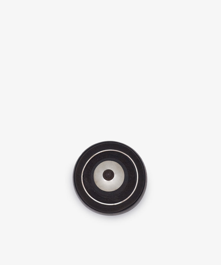 A round black and silver metal object with concentric circles, displayed on a white background, exudes the timeless elegance of Curiosity Box 17 from the Antje Weber collection.