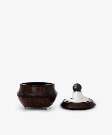 The Curiosity Box, a round African wood container with sterling silver accents, sits open next to its matching lid topped with a silver and dark spherical handle.
