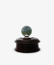 The Curiosity Box is a round African wood box featuring a metallic and green circular handle on top, displayed against a plain white background.
