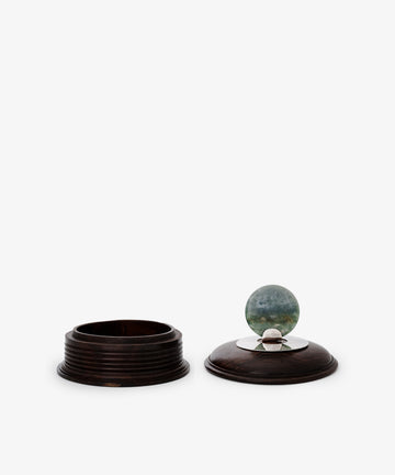 The Curiosity Box is a restored round vintage African wood container. Its removable lid features a metal accent topped with a greenish-blue marble, making it an eye-catching keepsake.