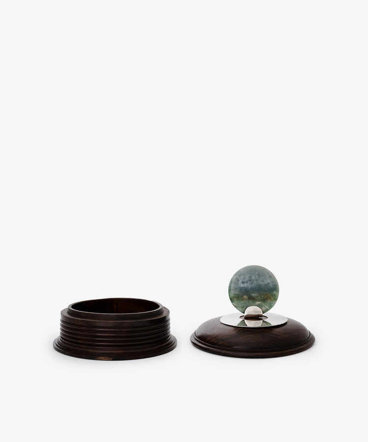 The Curiosity Box is a restored round vintage African wood container. Its removable lid features a metal accent topped with a greenish-blue marble, making it an eye-catching keepsake.