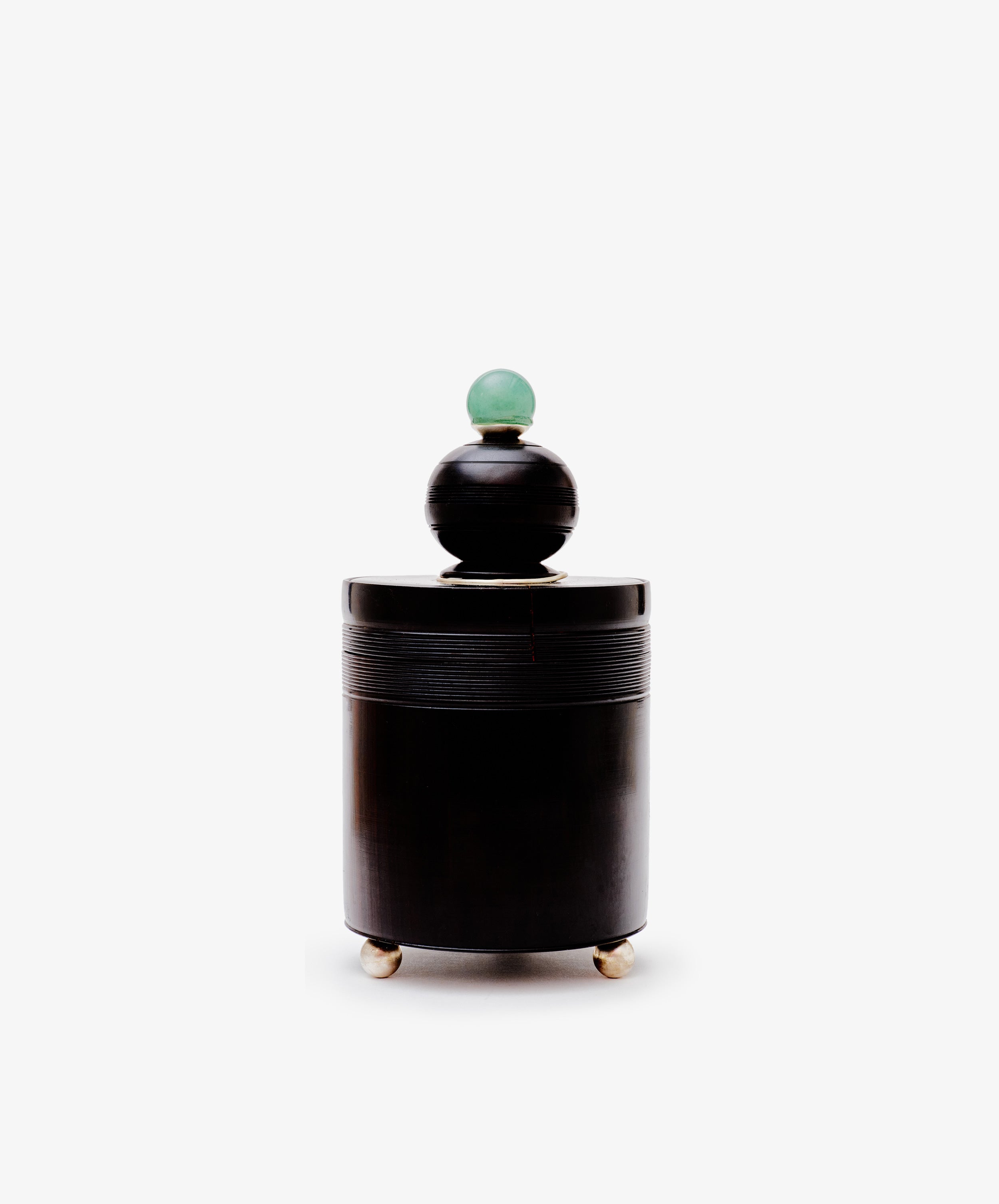 The Curiosity Box, a cylindrical black African wood container with a round lid topped by a green sphere, stands on three metallic feet in the style of Antje Weber art, inspired by vintage box designs against a white background.