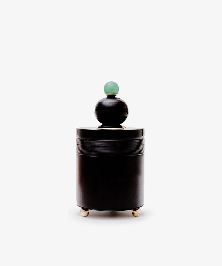 The Curiosity Box, a cylindrical black African wood container with a round lid topped by a green sphere, stands on three metallic feet in the style of Antje Weber art, inspired by vintage box designs against a white background.
