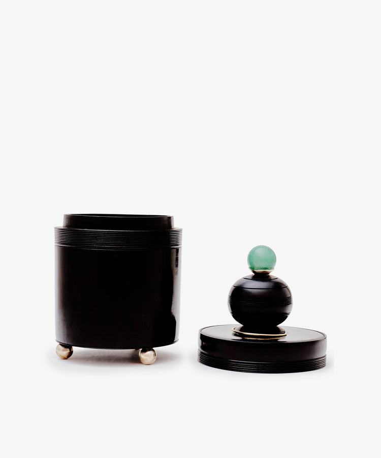 The Curiosity Box is a black cylindrical African wood box with three small metallic feet and a detached lid, featuring a round black handle topped with a green sphere, shown against a white background.
