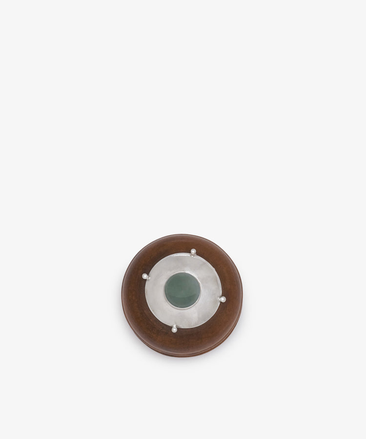 Curiosity Box 6: A round African wood box with a brown outer ring, silver metallic center, and green circular stone centerpiece, displayed on a white background.