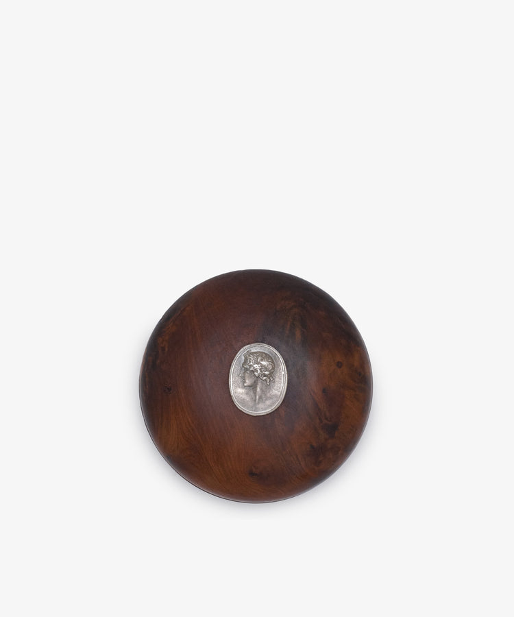 Curiosity Box 7: A round box crafted from dark African wood, featuring a small metallic oval medallion with a head profile embedded at its center.