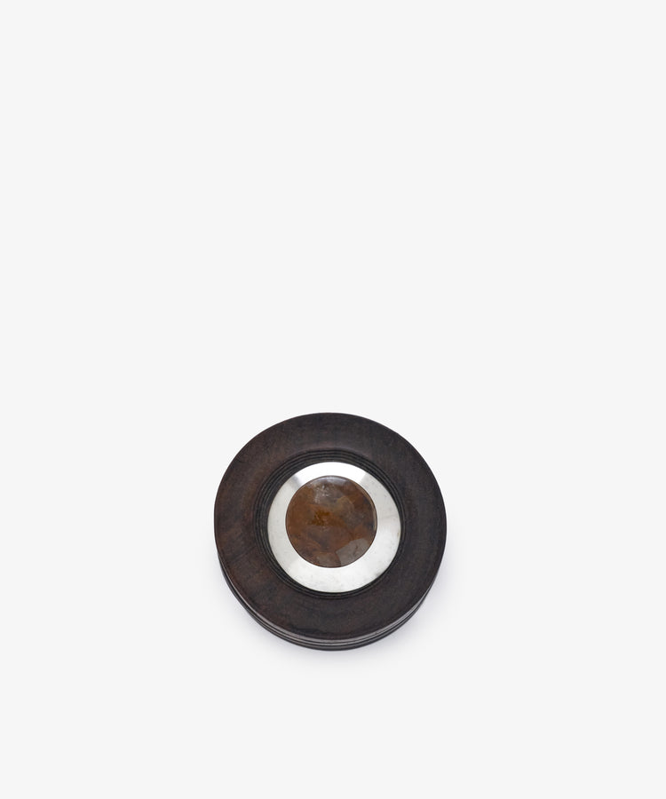 Curiosity Box 8: A round object with a dark outer ring, metallic inner ring, and brown center—resembling an African wood or vintage box—displayed on a white background.