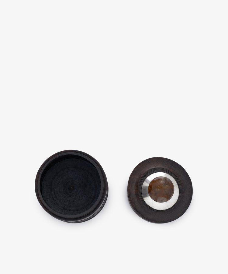 The Curiosity Box 8 is a round African wood box shown open, with its lid—featuring a striking metal and stone inlay at the center—placed to the right.